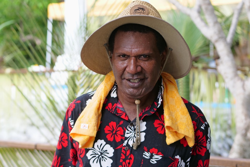 Man with hat and orange towel around neck wearing a floral shirt smiles at the camera.