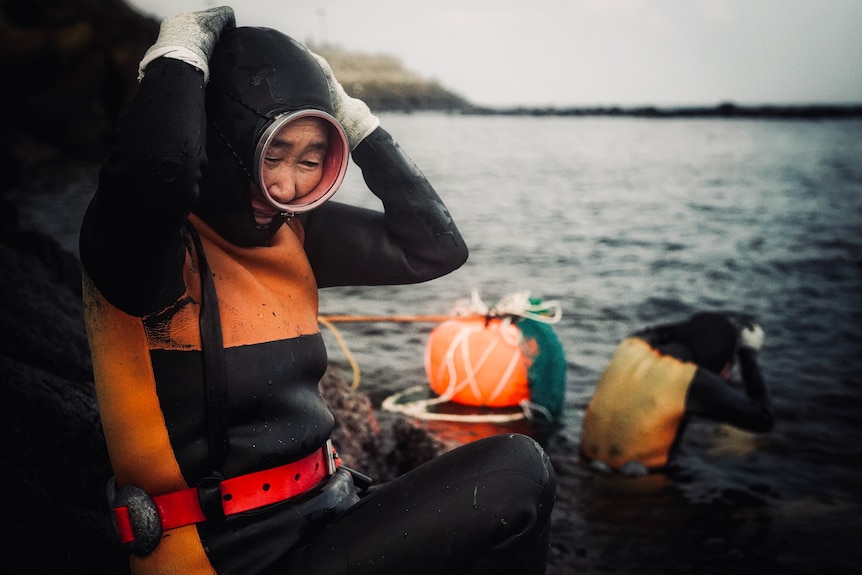 A woman holds her diving helmet on her head as she sits by the ocean in her diving gear.