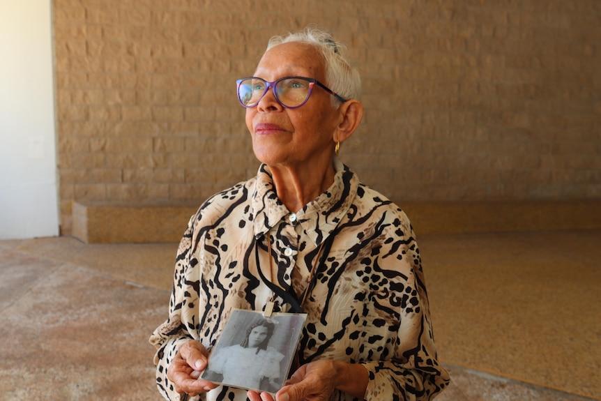 An Aboriginal woman holds a black and white photo of a girl and looks upwards