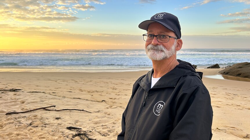 A man wearing a black hoodie and hat stands on the beach in front of the ocean.