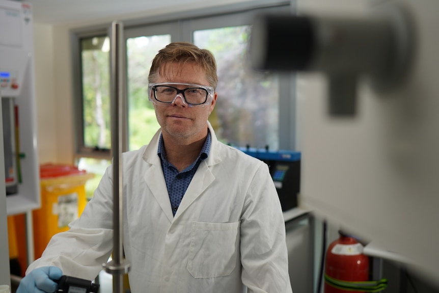 Man wearing a white lab coat and goggles standing in a laboratory room.