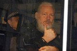 The side of a white police van shows three black rectangular windows with Julian Assange behind showing thumbs up.