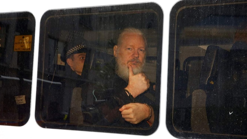 The side of a white police van shows three black rectangular windows with Julian Assange behind showing thumbs up.