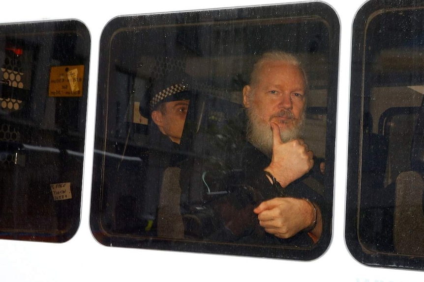 The side of a white police van shows three black rectangular windows with Julian Assange behind showing thumbs up.