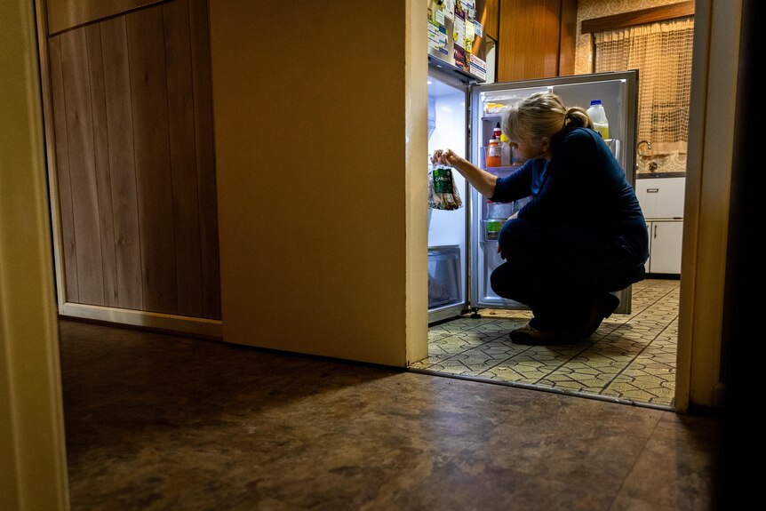 A woman crouched down in front of a fridge holding up a bag of rotten food.
