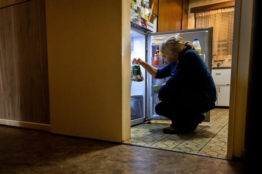 A woman crouched down in front of a fridge holding up a bag of rotten food.