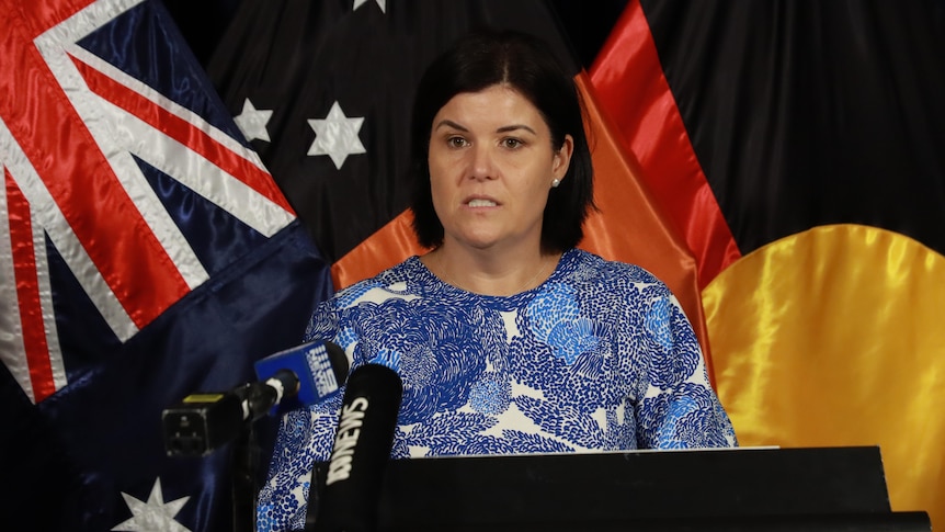 a woman with short dark hair speaking at a lecturn in front of australian flag