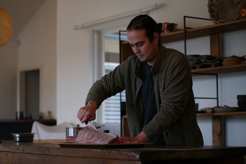 a man stands at a table holding a knife slicing into a chunk of venison meat