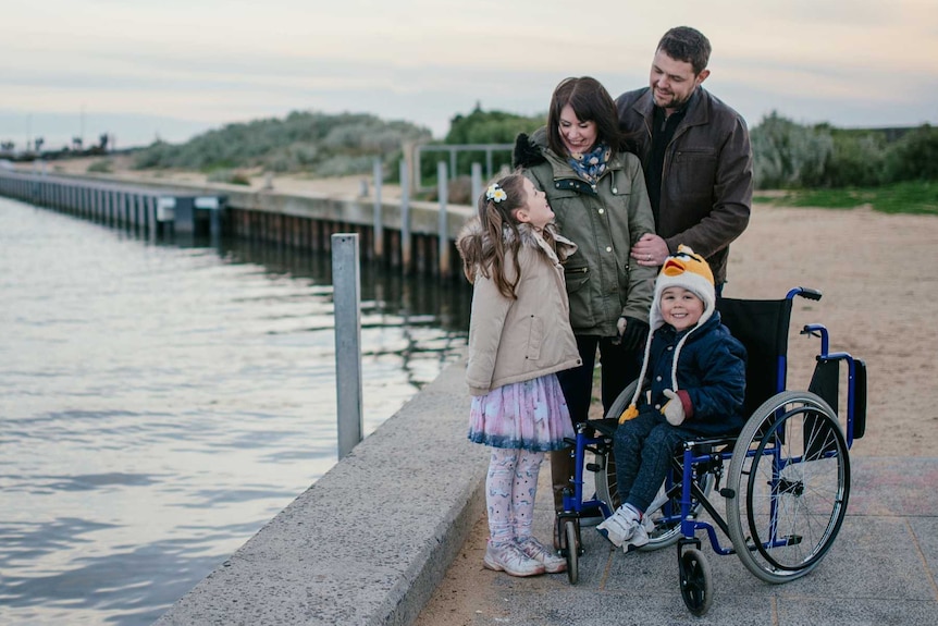 A man and a woman hold hands while walking alongside two children, one pushing the other in a wheelchair. They all smile.