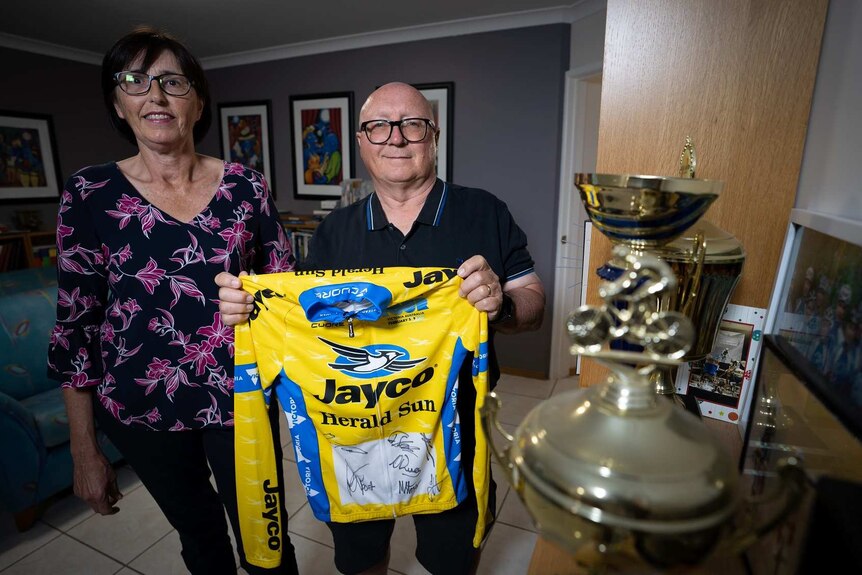 A smiling couple look at the camera holding their son's cycling jersey, standing next to trophies.