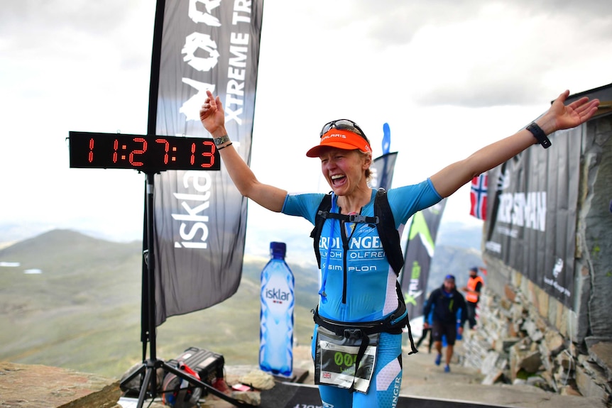 A triathlon runner screams in delight after crossing the finish line with mountains in the background