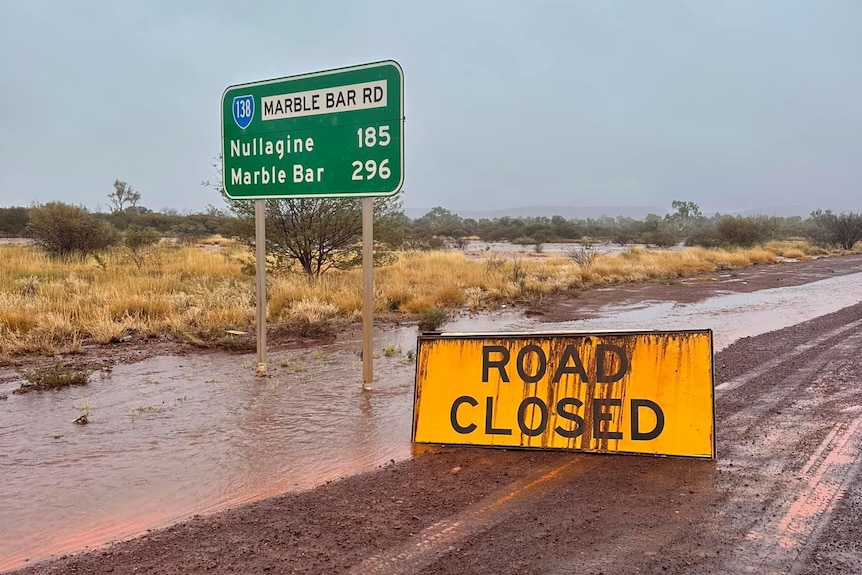 A muddy sign reads "Road Closed" in front of a sign with distances to Nullagine and Marble Bar on a muddy dirt road.