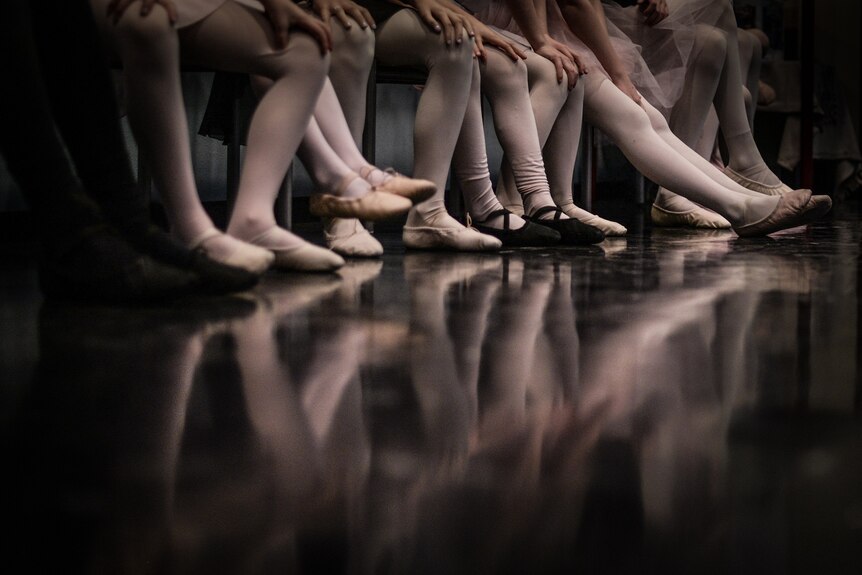 A photo of six young girls from the knees down sitting on chairs wearing ballet tights and shoes.