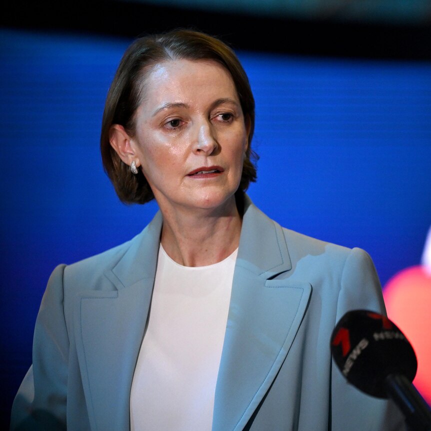 A woman with a brown bob wears a light blue blazer and speaks at a press conference, unsmiling.