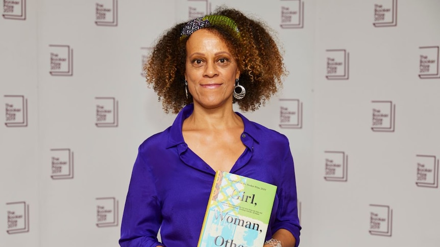 Bernardine Evaristo holds her book Girl, Woman, Other while standing in front of a promotional panel with the Booker logo on it