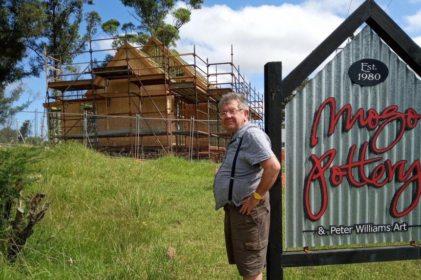 A man stands in front of half-built heritage church with scaffolding and the sign Mogo Pottery in the foreground.