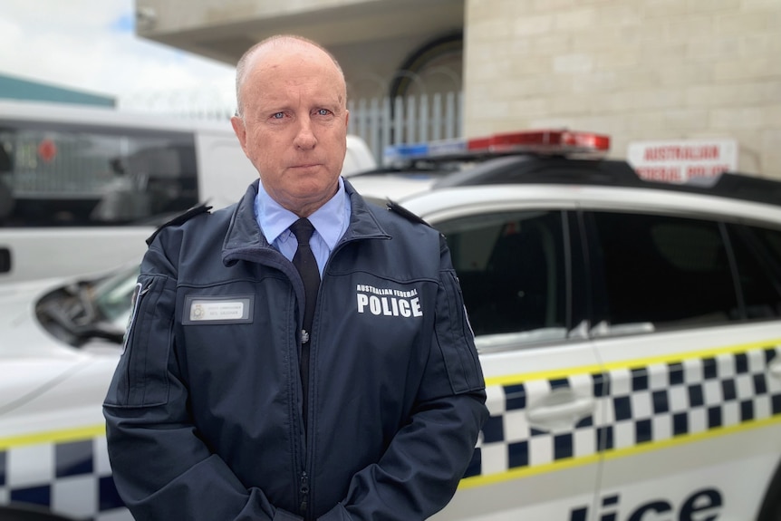 A man in police uniform looks sternly at the camera, standing in front of a marked police car.