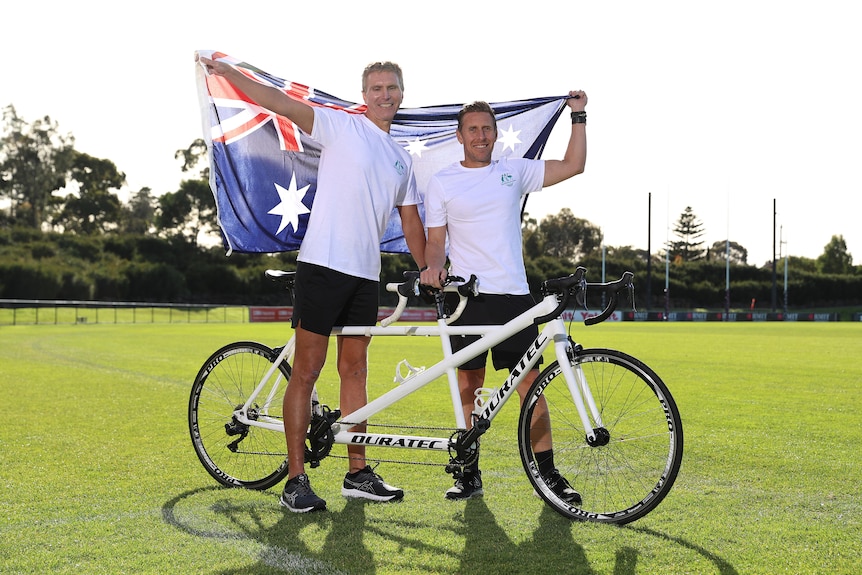 blind triathlete gerrard gosens and guide hayden armstrong hold an australian flag while standing either side of a tandem bike