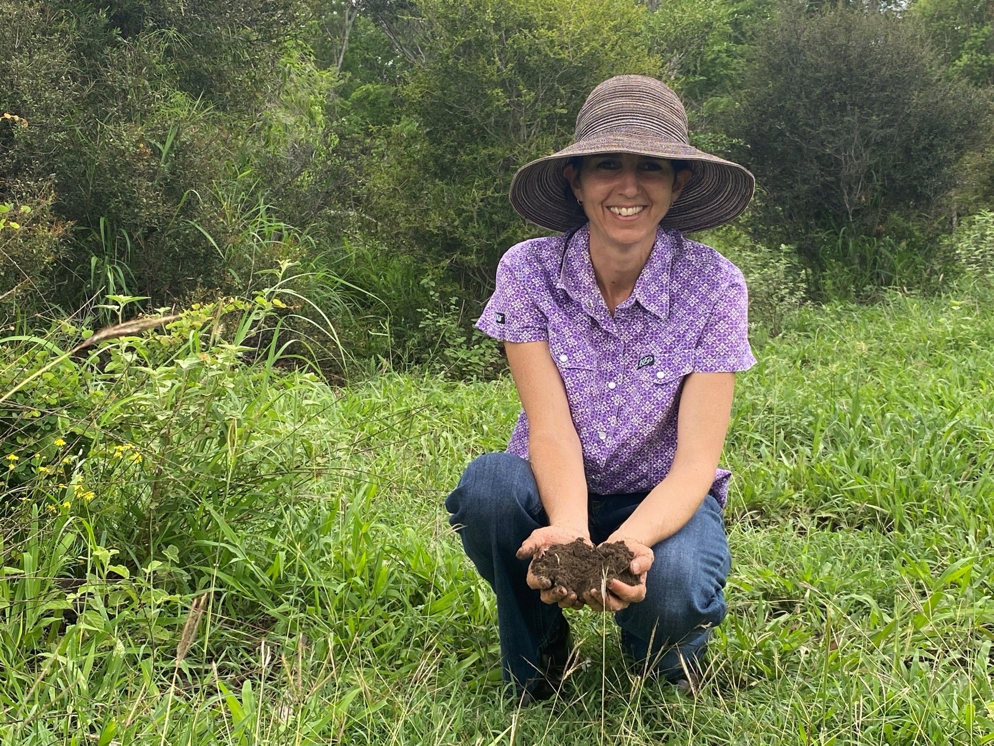 Uma mulher com um chapéu de abas largas está ajoelhada na grama e segura terra nas mãos.