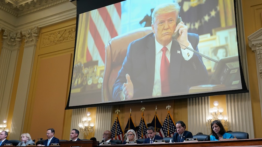 Donald Trump is shown on a big screen as people sit at formal table below him flanked by US flags