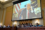 Donald Trump is shown on a big screen as people sit at formal table below him flanked by US flags