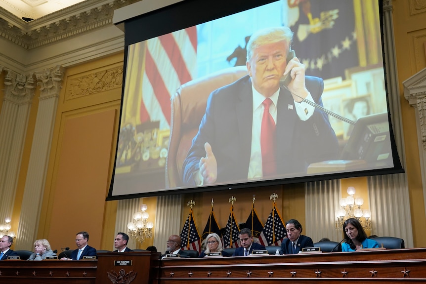Donald Trump apare pe un ecran mare în timp ce oamenii se așează la o masă oficială de sub el, înconjurați de steaguri americane