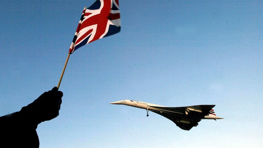 Looking up at a clear blue sky, a Union Jack flutters as a Concorde plane is seen flying in the distance