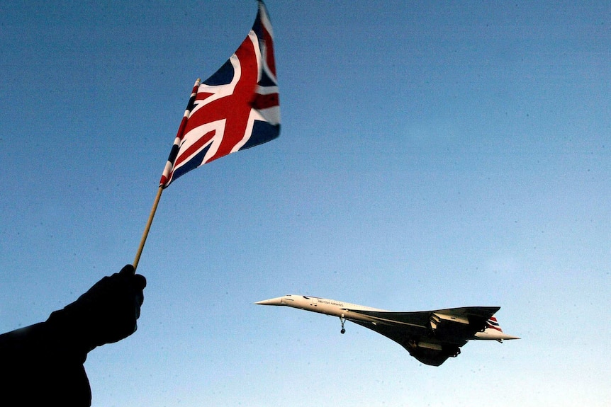 Looking up at a clear blue sky, a Union Jack flutters as a Concorde plane is seen flying in the distance