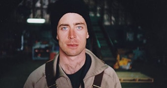 A man wearing work clothes and a black beanie, with bright blue eyes, stares towards the camera with a serious expression.