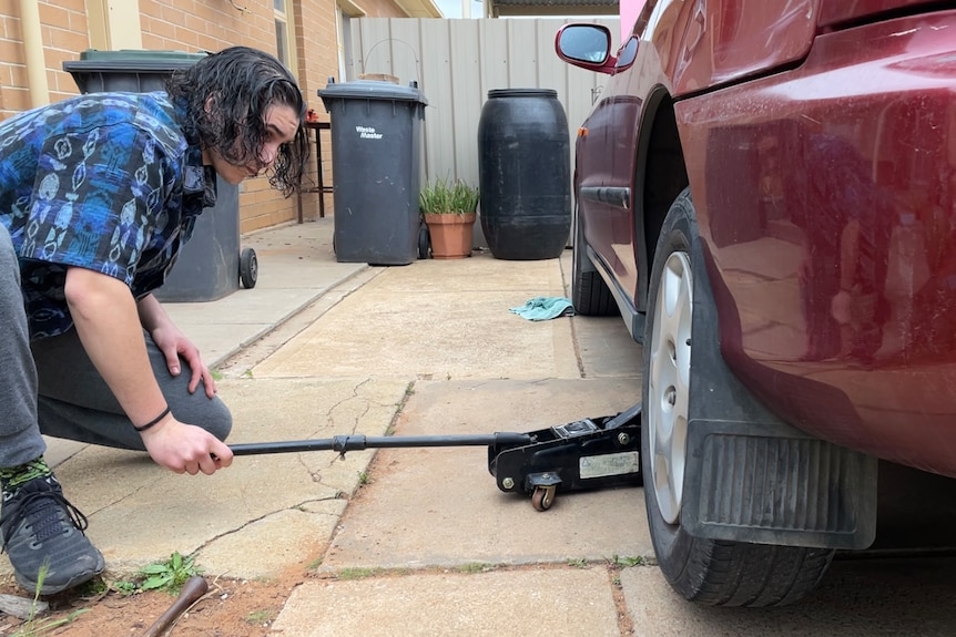 A man wears a blue shirt, grey pants and black shoes. He kneels down and jacks up a red car.