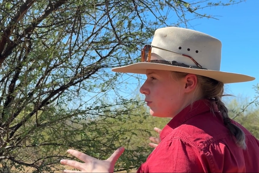 A woman wearing a red shirt and country hat standing in front of a green tree with thorns