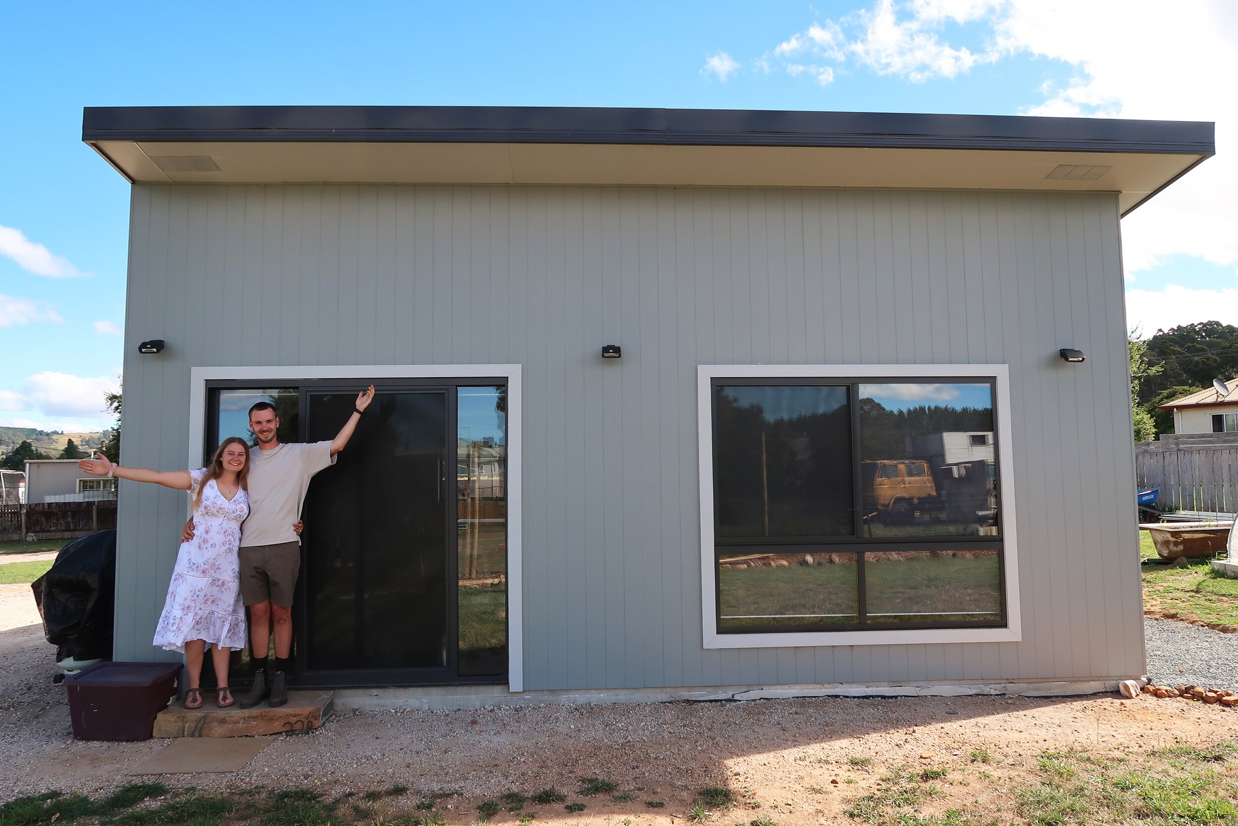 A young woman and a young man stand with arms oustretched in front of a small 