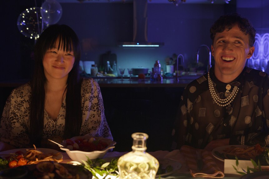 Young Asian woman with long dark hair sits at dinner table with white man with brown hair wearing dark shirt and pearls.