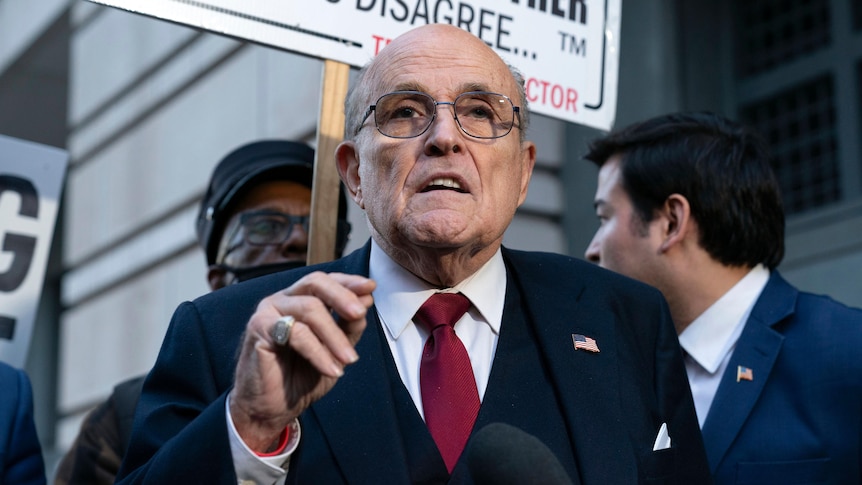 Rudy Giuliani speaks to the press, wearing glasses and a suit, hand raised to chest level