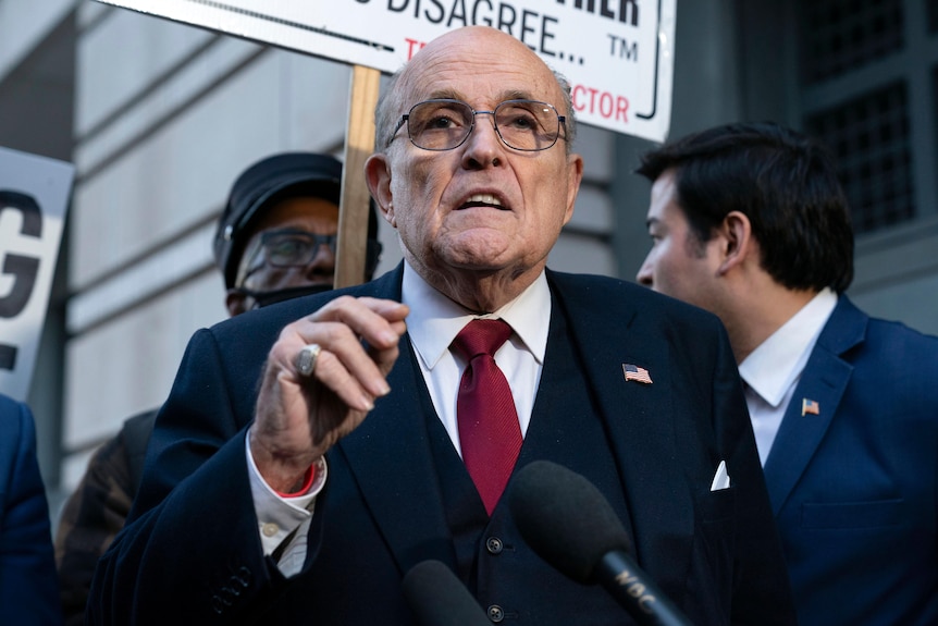 Rudy Giuliani speaks to the press, wearing glasses and a suit, hand raised to chest level