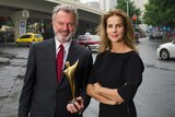 Actors Sam Neill and Rachel Griffiths standing on a street holding a trophy.
