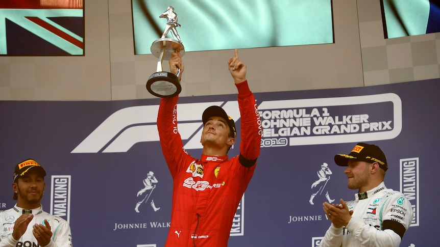 Charles Leclerc points to the sky holding a trophy in one hand. Lewis Hamilton and Valtteri Bottas applaud either side of him