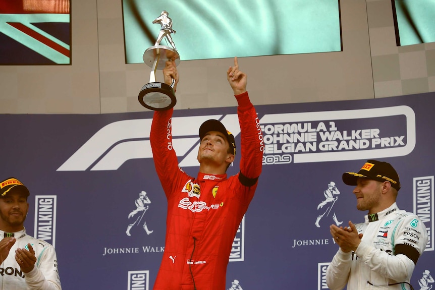 Charles Leclerc points to the sky holding a trophy in one hand. Lewis Hamilton and Valtteri Bottas applaud either side of him