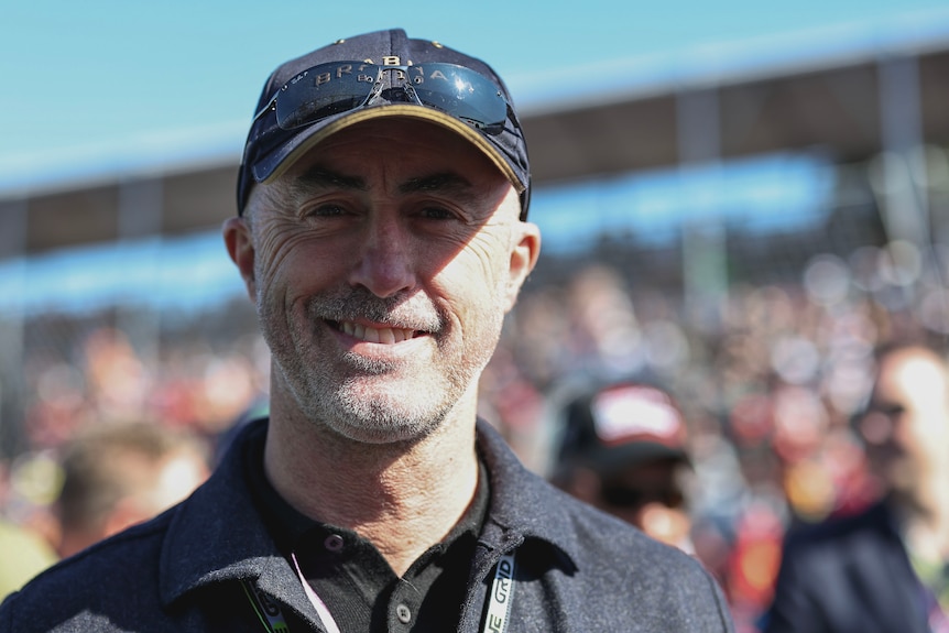 Former F1 driver David Brabham, wearing a black cap, smiling for a picture while standing on an F1 grid