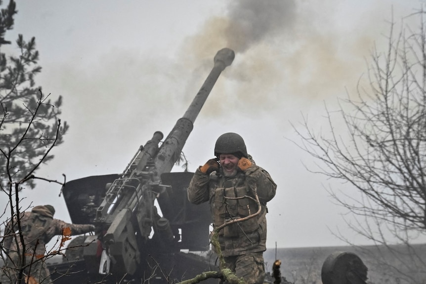 A uniformed man shouts and covers his ears as an enormous machine fires ammunition into the air