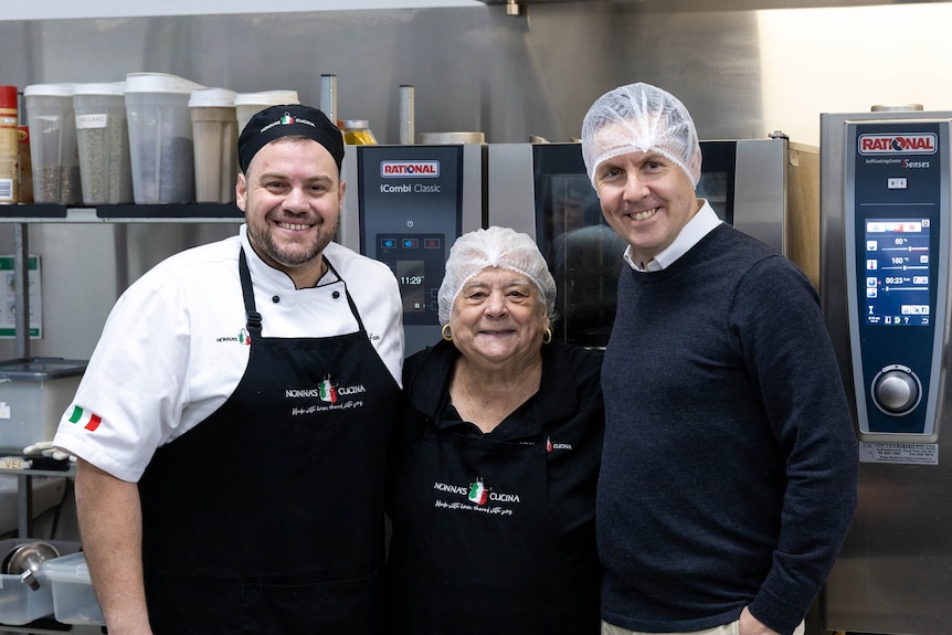 Two men stand either side of an older woman in a commercial kitchen