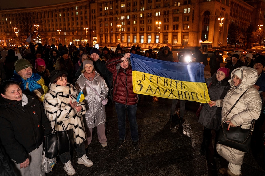People hold a Ukrainian flag during an outdoor protest at night among a crowd outside large buildings