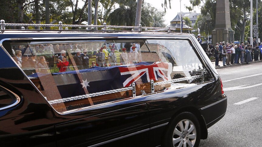 A close-up of a coffin draped in an Australian flag in a hearse driving as people watch on.