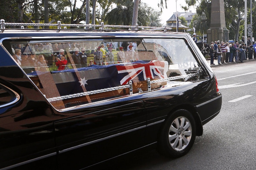 A close-up of a coffin draped in an Australian flag in a hearse driving as people watch on.