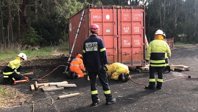 Emergency workers in high vis and hard hats gather around a red shipping container in a training scenario.