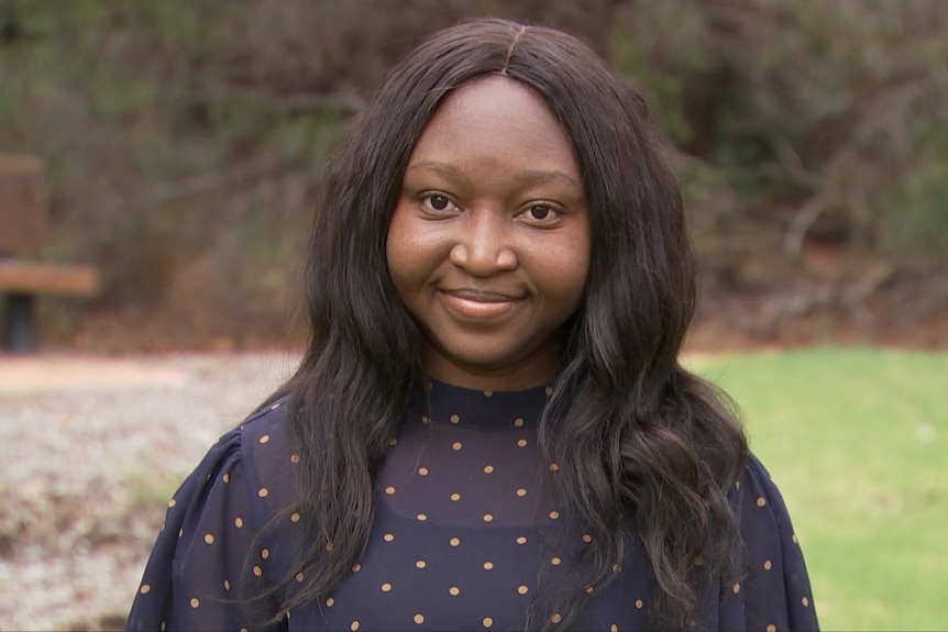 The professor is standing outside and smiling. She has long, black hair and deep brown skin, and wears a speckled dark blue top.