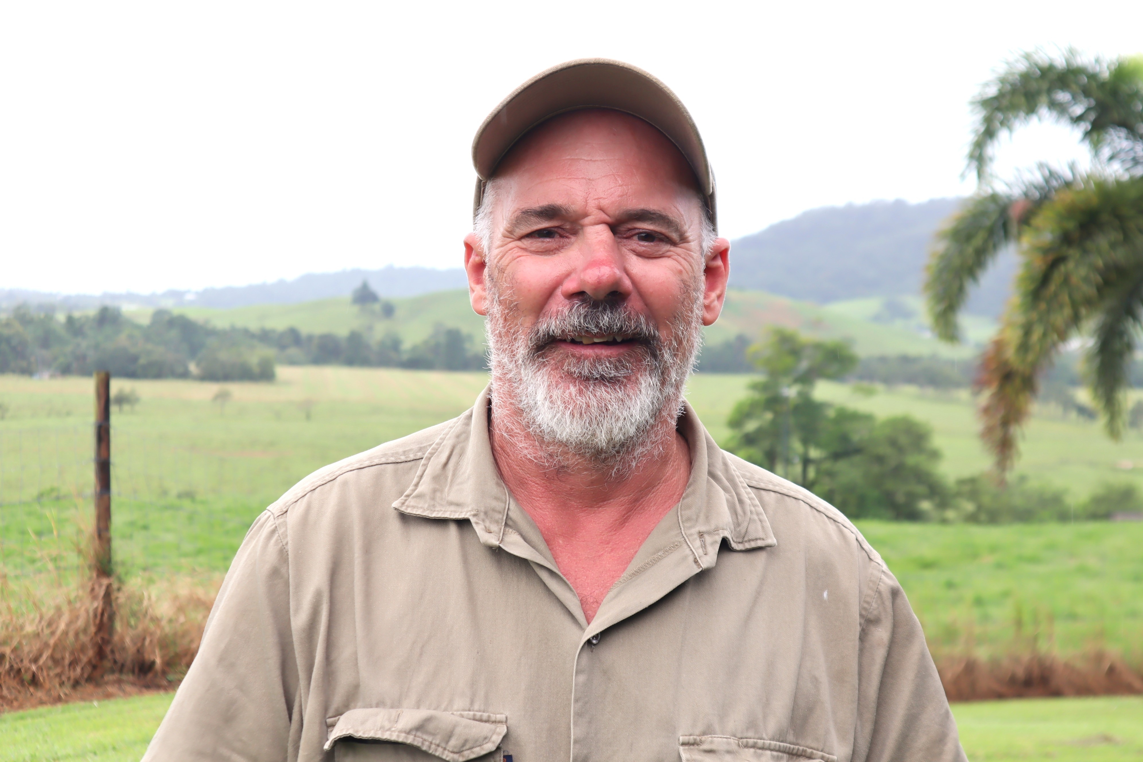 A man in a khaki shirt and cap with a beard stands in a green paddock.