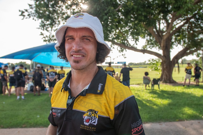 A man in a bucket hat wearing a club shirt smiles at the camera in front of an Aussie rules field