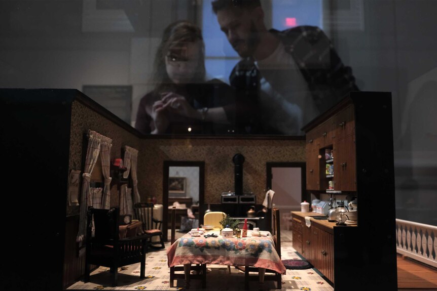 A blurred man and woman looking into a lit-up miniature living room, with dining table set with food and crockery.