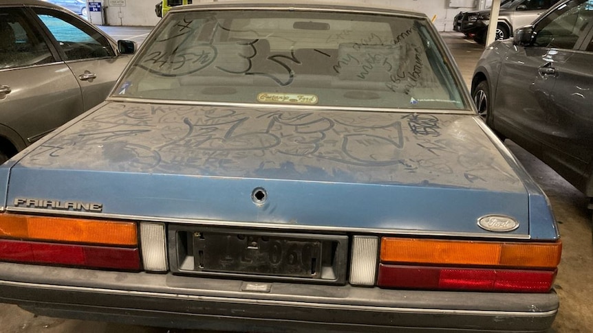 Blue car sits in a car park, covered in dust, people have written in the dust